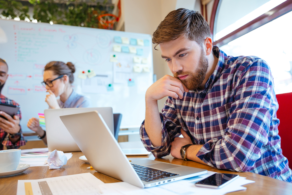 Concentrated bearded young man using laptop while his friends studying together